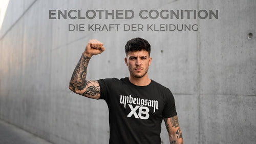 Junger Mann macht Sieger-Faust und trägt dabei ein schwarzes Premium Rock T-Shirt aus 100 % gekämmter Baumwolle mit der Aufschrift Unbeugsam. An der Wand im Hintergrund steht geschrieben: Enclothed Cognition - Die Kraft der Kleidung.