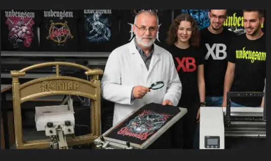 Prüfer schaut sich mit Lupe Druckverfahren an und wird dabei von jungen Leuten die jeweils ein Premium Rock T-Shirt von XB tragen beobachtet.