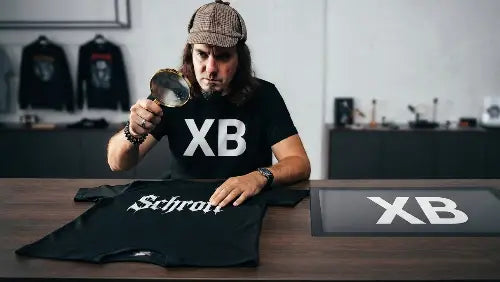 Mann vor Schreibtisch trägt ein Premium Rock T-Shirt von XB & untersucht mit der Lupe ein Billig-T-Shirt (Schrott).