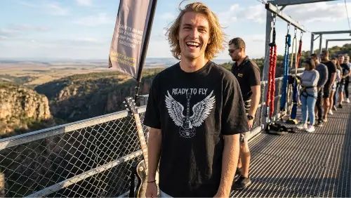Junger Mann auf Bungee Jumping Anlage trägt schwarzes Premium Rock T-Shirt aus 100 % Bio-Baumwolle mit komplexem Flügel-Gitarren Design aus Öko-Tex zertifizierten Farben in einer Grammatur von 240g/m².