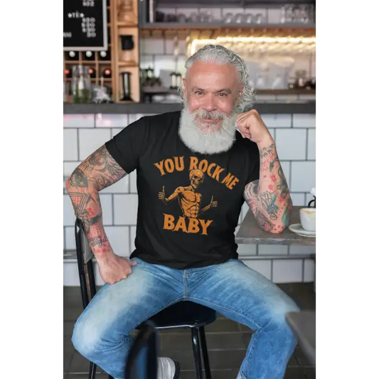 Mann sitzt im Bistro und trägt ein Rock T-Shirt für Männer mit hochwertigem Druck, you rock me baby und einem Skull der die Daumen nach oben hält.